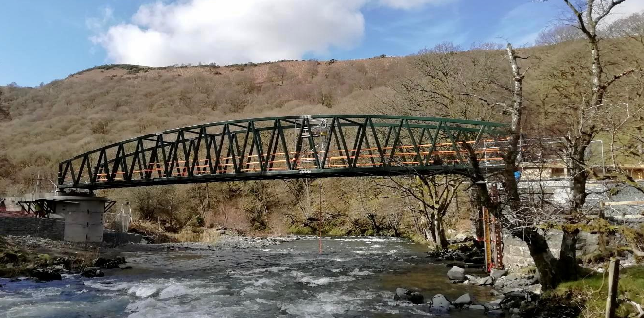 A scenic view of a permanent bridge over a river, framed by a towering mountain in the distance.