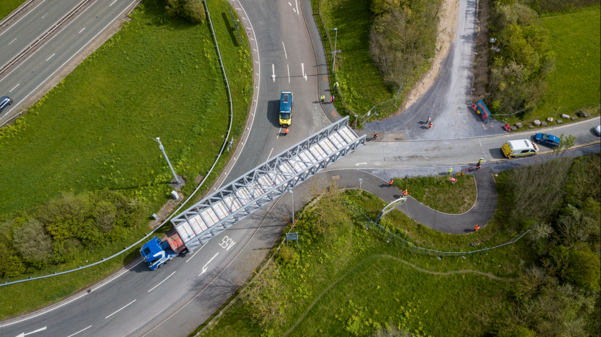 Aerial view of a Beaver Bridges truck transporting a massive steel truss section across a road junction, supported by an escort vehicle.