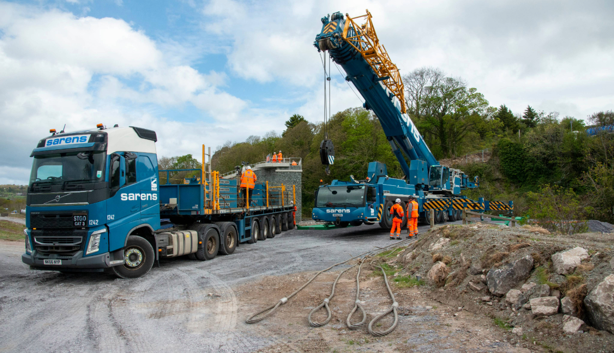 A blue Sarens crane and Volvo heavy-haulage truck on a gravel construction site, prepared for a Beaver Bridges installation project.