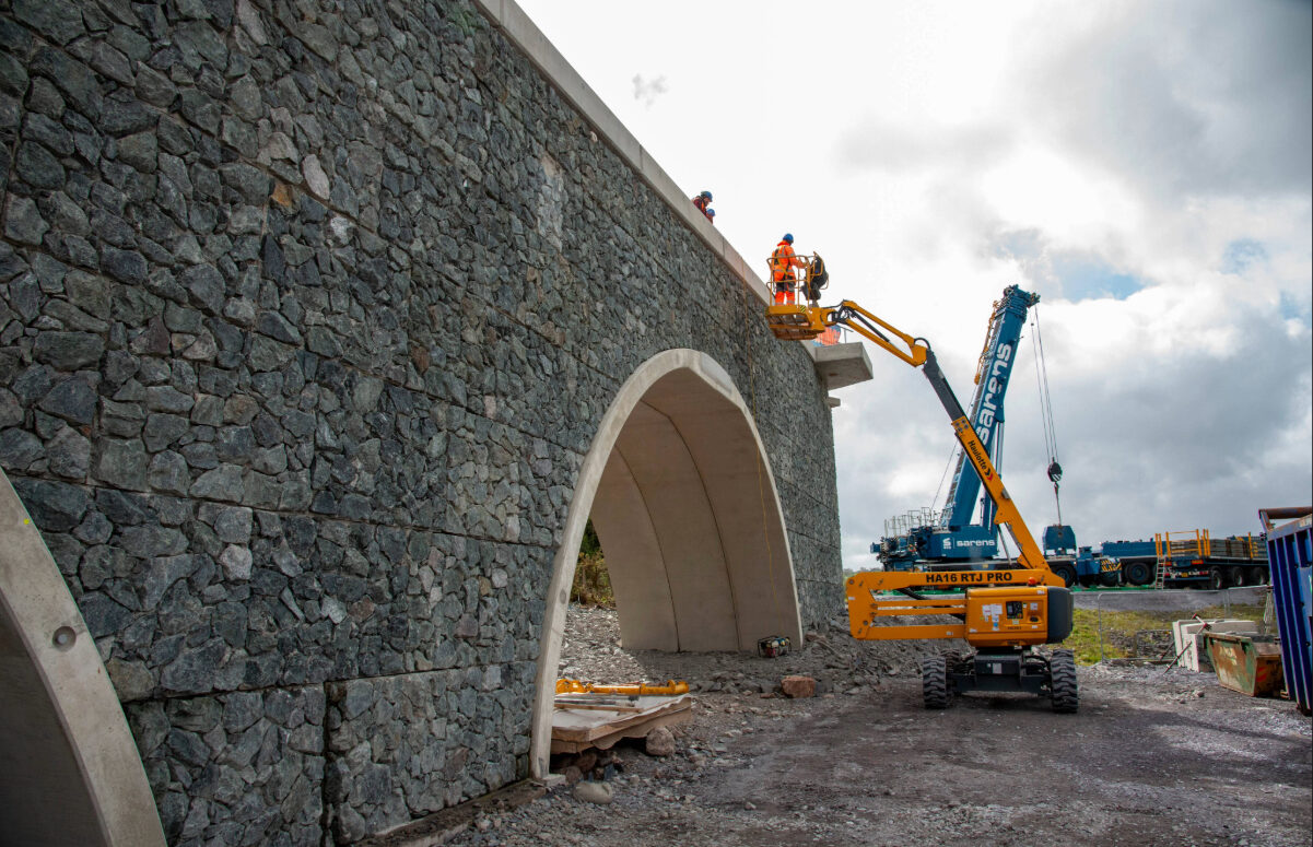 Workers on a cherry picker finalise a stone-clad arch bridge, with a large Sarens mobile crane and support equipment in the background
