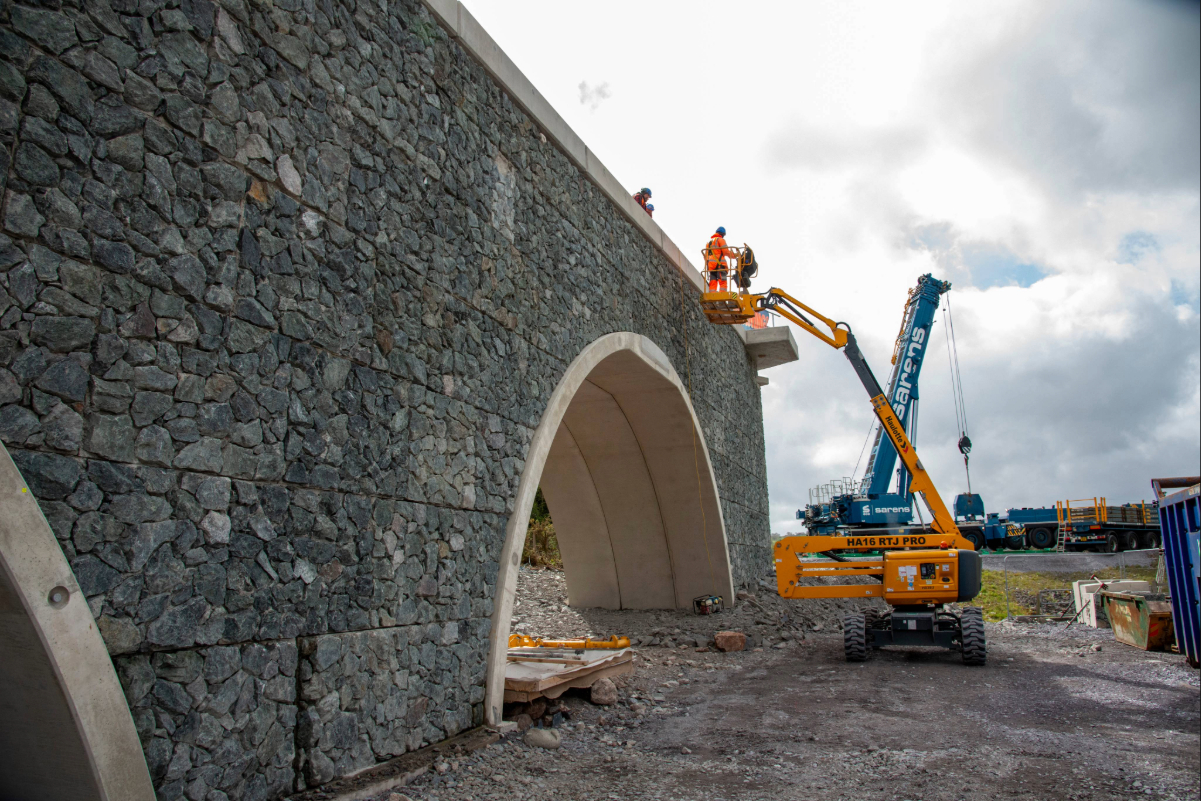 Workers on a cherry picker finalise a stone-clad arch bridge, with a large Sarens mobile crane and support equipment in the background