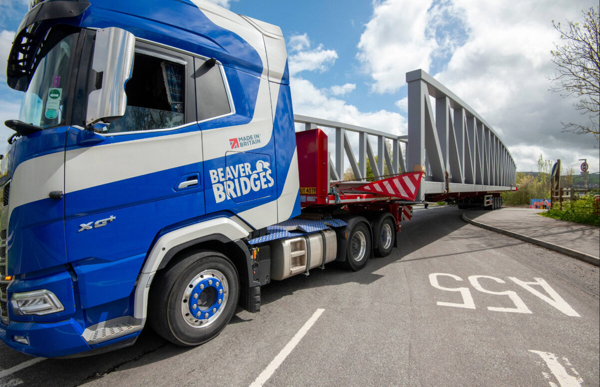 A blue and white branded Beaver Bridges truck carrying a large grey steel bridge section on a trailer, showcasing British manufacturing.