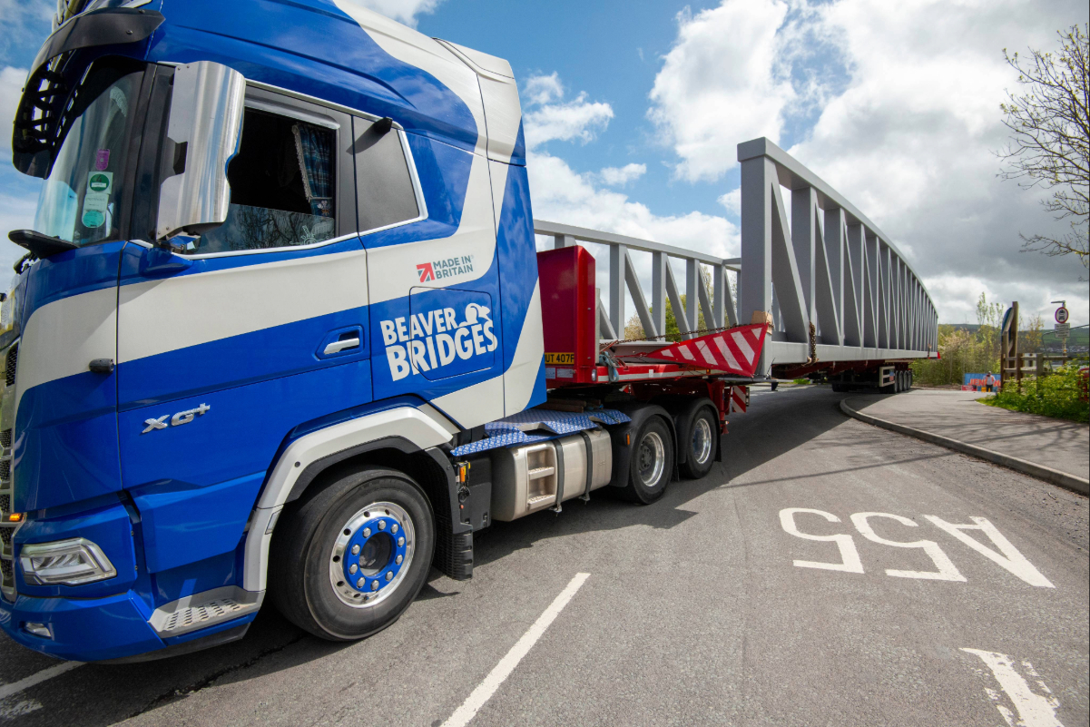 A blue and white branded Beaver Bridges truck carrying a large grey steel bridge section on a trailer, showcasing British manufacturing.