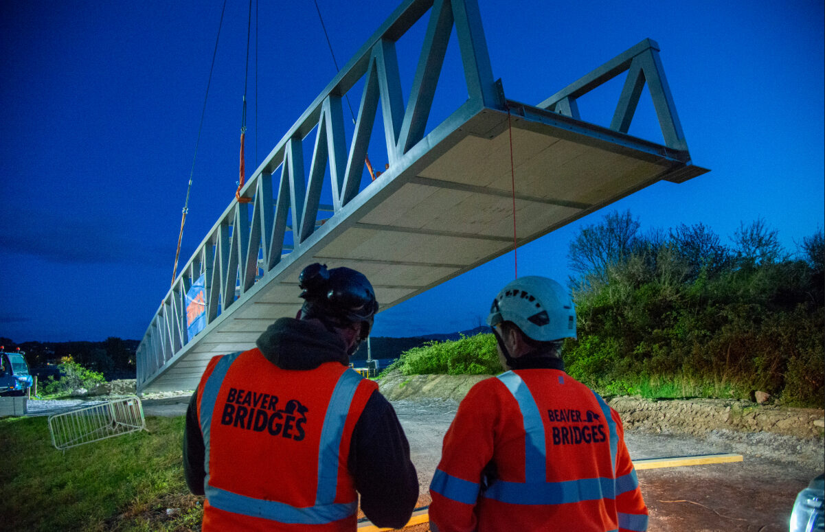 Two Beaver Bridges engineers in orange hi-vis vests watch as a large steel bridge section is lifted by crane during a night installation.