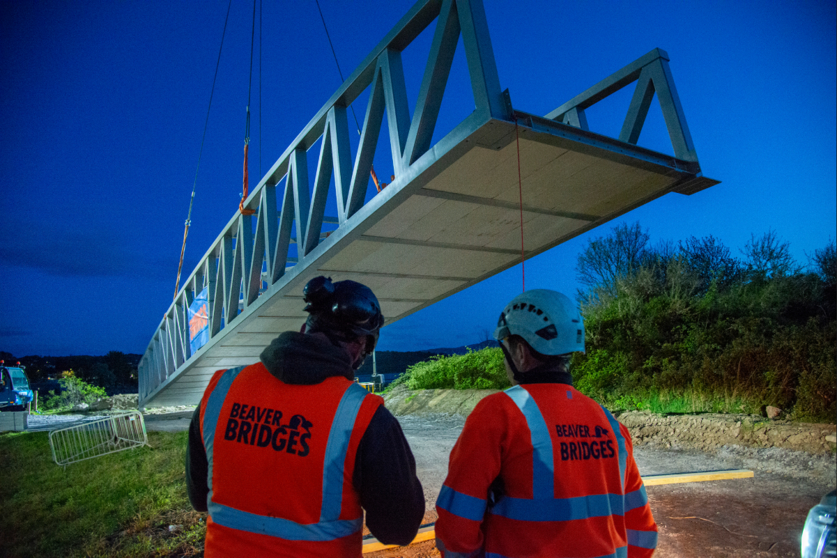 Two Beaver Bridges engineers in orange hi-vis vests watch as a large steel bridge section is lifted by crane during a night installation.
