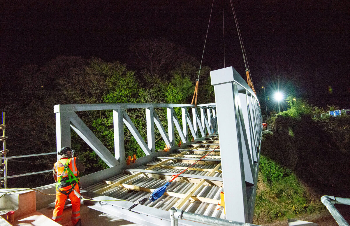 A Beaver Bridges engineer in safety harness monitors a steel truss bridge being lowered into place by crane during a nighttime installation.