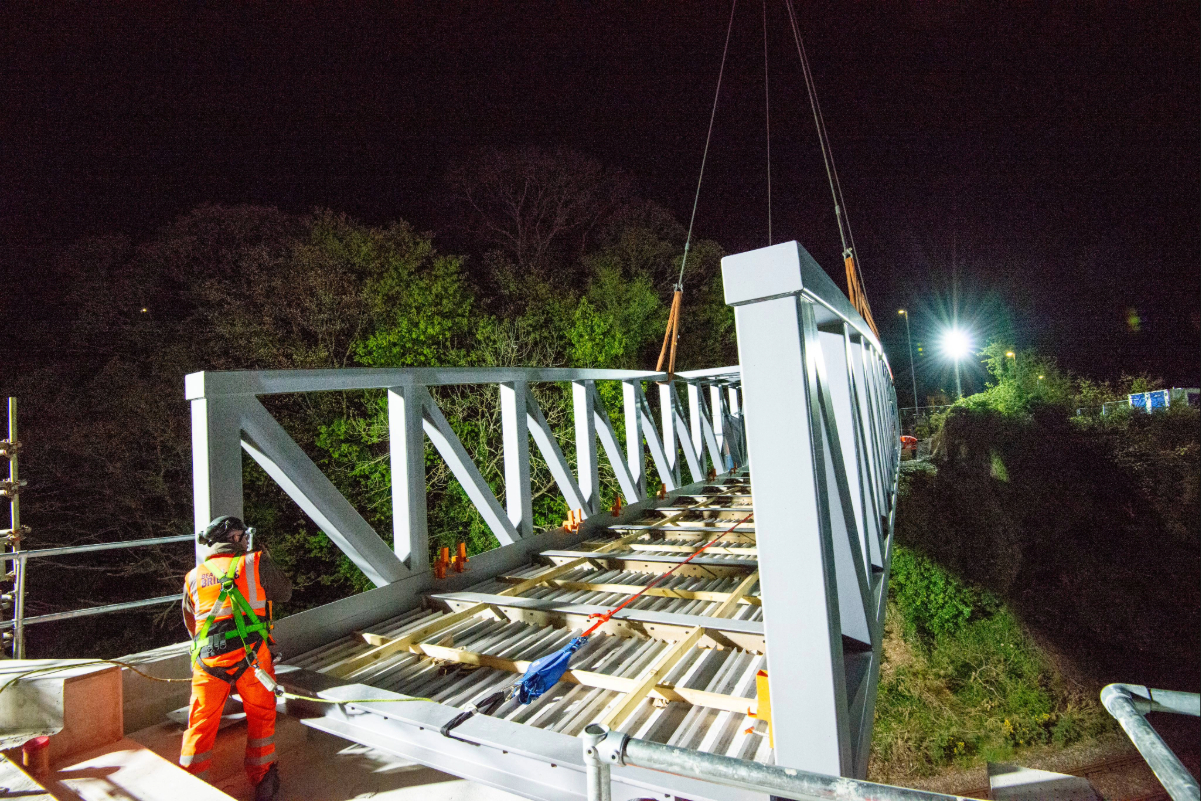 A Beaver Bridges engineer in safety harness monitors a steel truss bridge being lowered into place by crane during a nighttime installation.